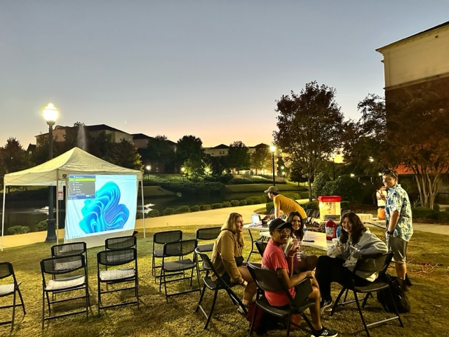 students sit outside on the quad in chairs and watch a movie projected onto a canvas